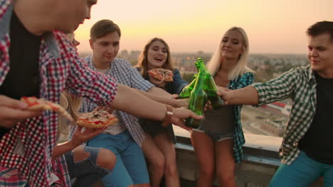 Young-girls-and-boys-clinks-glasses-and-drinks-beer-from-green-bottels-on-the-party-with-friends-on-the-roof-at-the-sunset.They-are-sitting-together-eat-hot-pizza-after-in-summer-everning.